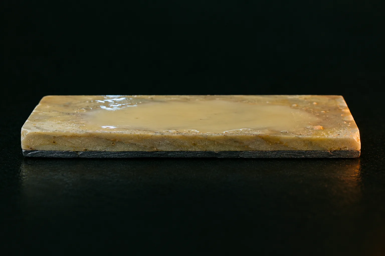 Une pierre à aiguiser rectangulaire vue de trois quarts, sa face supérieure d’un jaune crème couverte d’une boue laiteuse jaune qui s’accumule au centre, et une couche d’ardoise gris sombre collée sous toute sa longueur