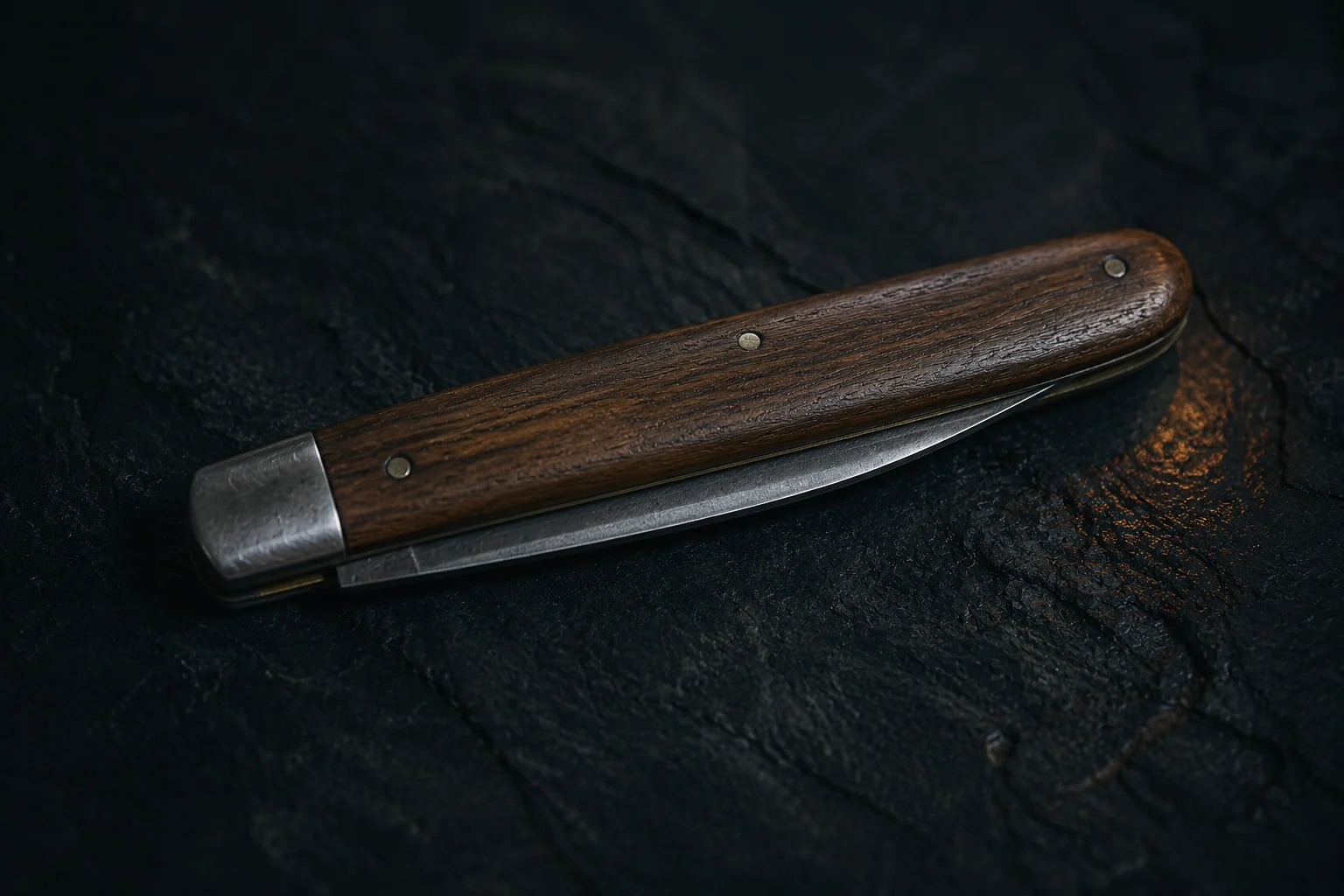 Un couteau de poche pliant fermé, à plaquettes de bois brun et mitre d’acier, posé à plat sur une ardoise sombre, le dos de la lame affleurant le long du manche