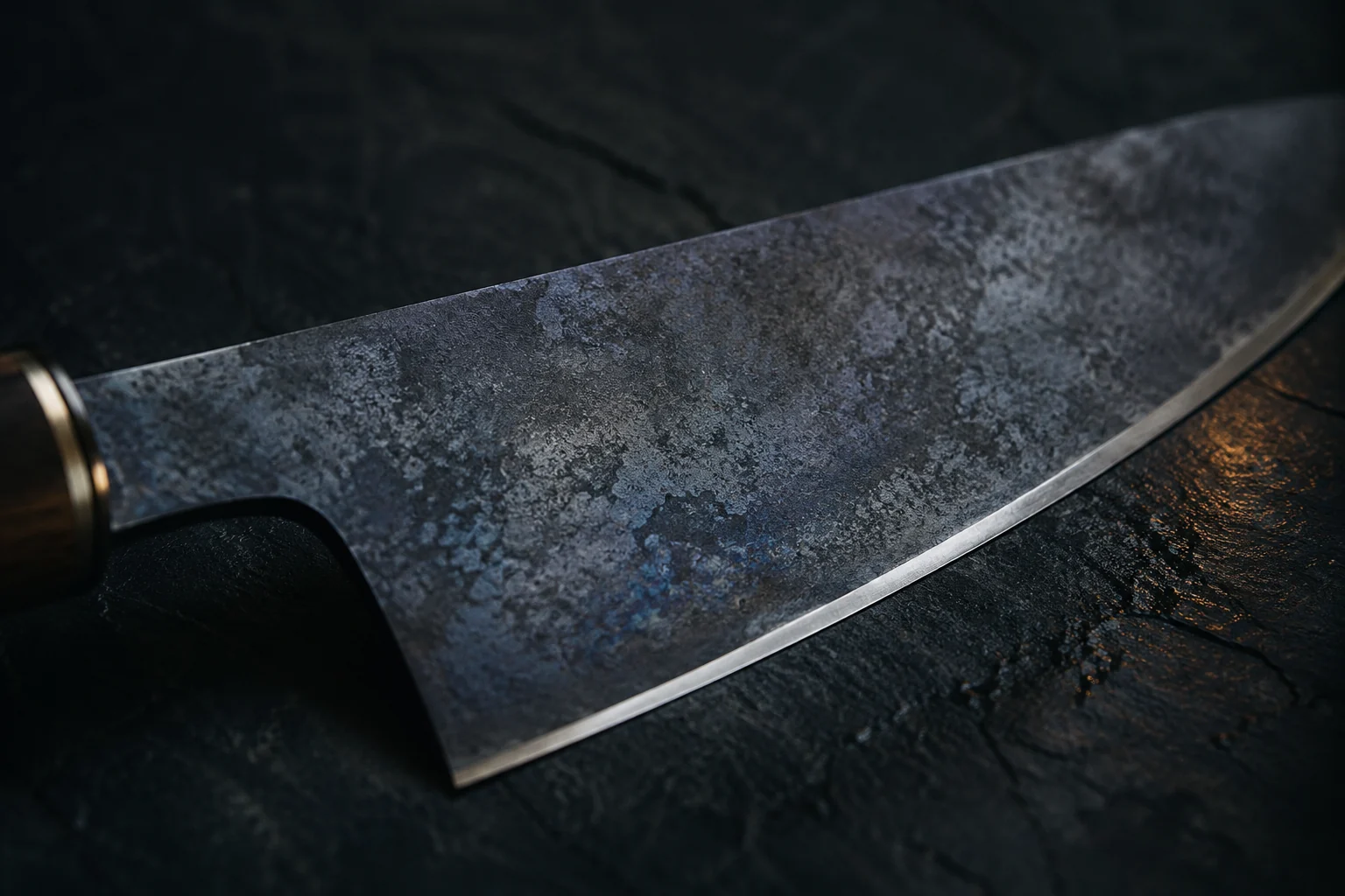 Gros plan sur la lame d’un couteau de cuisine entièrement recouverte d’une patine gris-bleu marbrée, avec des reflets violacés, sans le moindre point de rouille orange, et une bande d’acier restée claire le long du fil